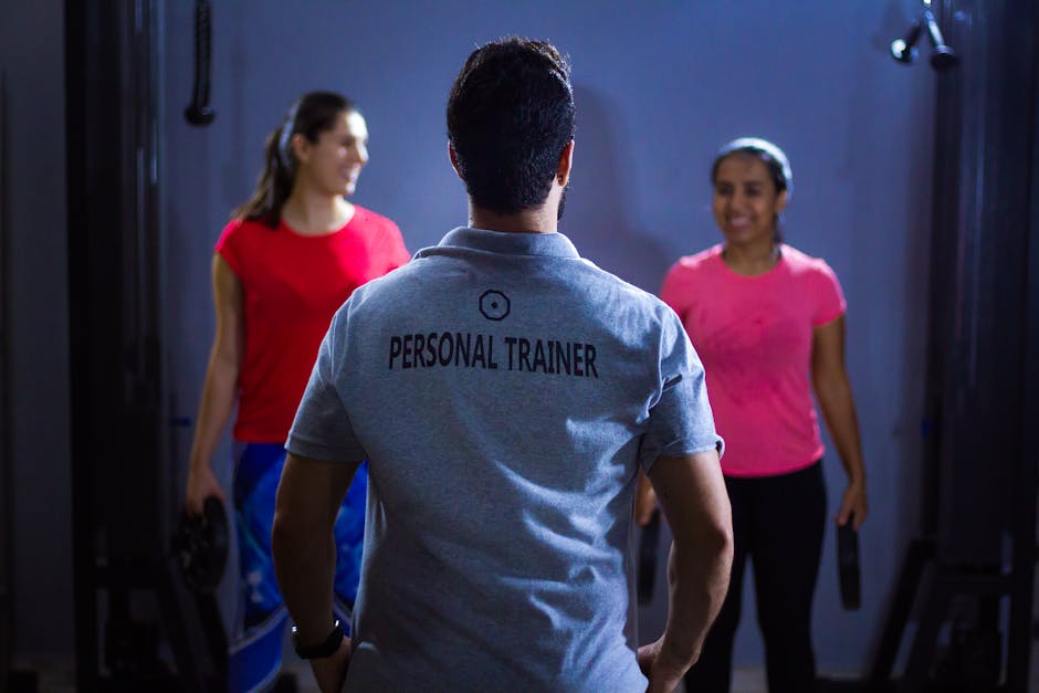 Back view of a personal trainer guiding two women during a gym session indoors.